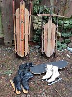 Photo of two vintage wooden sleds standing up and three pairs of ice skates with a King Kong skateboard placed in front.