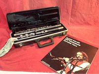 Flute disassembled and stored in open hard black case beside the music book on a red surface.