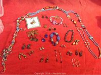 Photo showing assorted pairs of earrings, beaded necklaces, bracelets and brooches on red cloth background.