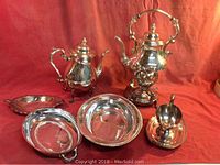 Seven silver plated service pieces arranged on a red cloth backdrop showing a coffee pot on a warming stand with sterno container, a teapot, a creamer, and four trays of various shapes and sizes