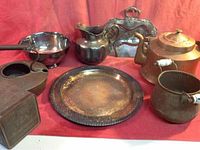 Photo showing assortment of metal pots, bowls, a copper kettle, silver plated platter and bowl, brass pots, and decorative tray, all on red cloth.