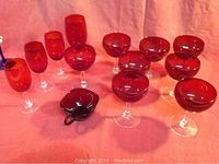 Photo showing 6 red champagne glasses with clear stems, 4 red wine glasses with clear stems, and 1 red cup, all placed on a pink cloth background.