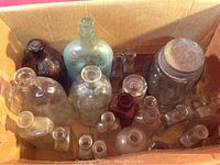 Photo of an open cardboard box containing a variety of old dug-up beach bottles in different sizes, shapes, and glass colors including clear, blue, and brown.