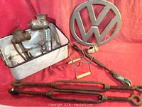Photo showing two metal meat grinders mounted on painted enamel pan, vintage VW Bus insignia, two wooden-handled chest grips, and two large rusted metal turnbuckles.