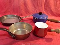 Photo showing a blue cruet sauce pan with lid and wooden handle, red cruet sauce pot with wooden handle, copper pot, and crepe pan.