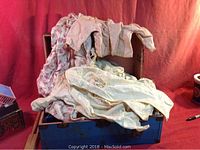 Small blue metal trunk open with vintage doll clothes draped over and inside it, items appear aged with pastel and floral patterns