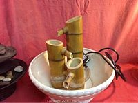 White ceramic bowl fountain with bamboo-style columns and attached power cord, placed against red fabric background.