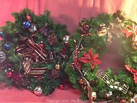 Photo showing three decorated green wreaths with red, gold, and blue ornaments, pine cones, ribbons, and other holiday decor elements.
