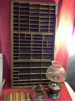 Old wood shadow box in dark brown with multiple small compartments for miniatures, placed vertically on a red cloth backdrop, accompanied by two oil lamps with glass chimneys and metal bases.