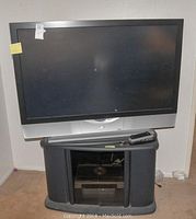 JVC flat-screen TV on top of black TV stand with glass cabinet doors, remote controls visible on stand.
