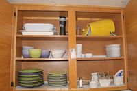 Wooden cabinet with shelves containing various dishware including plates, bowls, cups, salt and pepper shakers, and a yellow beverage dispenser.