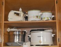 Photo showing kitchen shelf with white Hamilton Beach hand mixer, two mini crock pots with vegetable pattern, silver frying pan lid, stainless steel strainers, and large CorningWare crock pot with lid.