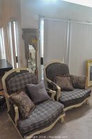 Two upholstered armchairs side by side with pillows, and carved wood floor mirror behind