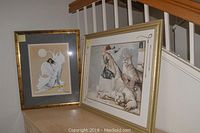 Both framed artworks placed side by side on a cabinet against a stair railing, one larger with woman and tigers, other smaller with abstract man and moon.