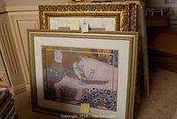 Photo showing framed reclining woman print with detailed gold and blue accents leaning against a wall along with other framed art.