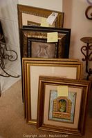 Four framed artworks stacked leaning against wall. Frames are gold and wood tones. Visible artwork includes classical urn in tiled niche and another with ornate gold frame.