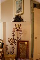 Photo showing two wrought iron wall sconces placed against the wall, a framed picture hanging above, and other frames leaning nearby.