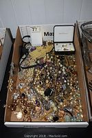 Wide shot showing a large assortment of costume jewelry including necklaces, bracelets, brooches, pins, and earrings in a cardboard box.