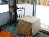 Photo showing two metal plant stands with bottom shelves and a beige twill fabric covered ottoman next to a sliding glass door, with visible wear on plant stands and staining on ottoman