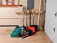 Wide view of the entire lot of yard and gardening tools arranged near garage wall, showing snow shovels, rakes, manual aerator, watering can, hose, trimmer, and other tools.