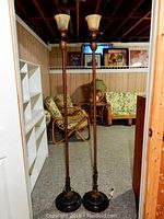 Full view of two tall, metal floor lamps with round bases and pineapple decorations at the mid-section, topped with opaque upward-facing glass shades in a basement setting.