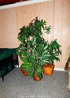 Three faux plants in woven baskets placed against a wood-paneled wall on carpeted floor.