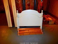 Photo of white solid wood headboard and footboard with natural wood slats rolled beneath, placed against a wall on a concrete floor.