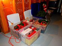Six large plastic tubs filled with Christmas decorations including ornaments, greenery, and boxed items, arranged on a basement floor.