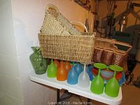 Woven wicker picnic baskets, colored non-breakable wine glasses arranged on a white table with plates and bowls in the background