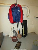 Wide view showing NFL Giants jacket hanging on wall, Dunlop golf bag standing beside two wooden baseball bats, two footballs, one basketball on the floor with two pickleball paddles.