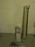 Two heaters side by side against a wall: Lakewood portable radiator style heater with visible control knob and a tall, slim electric baseboard heater.