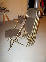 Four metal folding chairs, one open and three folded, showing gold metal frames and vinyl seats/backs.