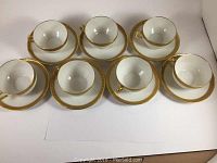 Top-down view showing all seven matching tea cups seated on their saucers, revealing consistent double gold encrusted borders on white porcelain.