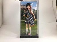 Front view of Barbie doll in traditional Scottish attire inside original packaging, holding bagpipes with a scenic Scottish background.
