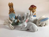 Photo showing nine assorted bunny figurines in various poses and styles including porcelain and ceramic pieces, some with paint wear.