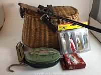 Full view of lot including fly fishing rod with reel, wicker and leather creel, fishing lure pack, decorative fish tin, and fishing scale with hook.