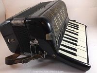 Side view of vintage accordion showing black finish, bellows, and leather strap with some wear and peeled spots.