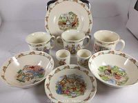 Full set of Royal Doulton Bunnykins items arranged showing all pieces: one plate, three mugs, two bowls, two egg cups, one saucer, with clear bunny illustrations and decorative border.