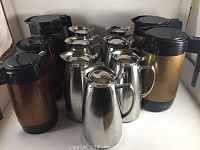 Overall group of 12 thermal jugs including five plastic and seven stainless steel arranged closely showing variety in color and material.