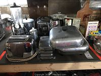 Collection of retro kitchen appliances on wooden table including stainless steel toasters with braided cords, metal pots, pans, glass blender, and metal coffee pot.