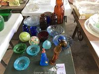Full display of assorted colored glassware and decorative glass items on table including bowls, vases, bottles and figurines in blue, green, amber and multicolored glass.