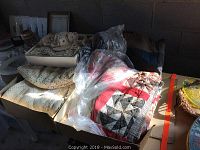 Two quilts stacked with one wrapped in plastic showing a red and gray circular star design. Multiple boxes hold hand crocheted lace and doilies, cream and natural yarn colors visible.