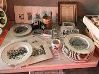 Photo showing 12 matching plates stacked and spread flat, black and white illustrations of historic Sandy Spring architecture, 4 mugs, and other small boxed items on a table.