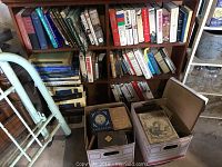 Books on wooden shelves and two boxes on the floor filled with various books. Books vary from newer to older editions.