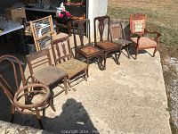 Photo of seven old wooden chairs and one vintage folding chair arranged outdoors showing variety of styles and wear.