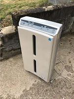 White Atmosphere air purifier with multiple control knobs and buttons on top front panel shown outdoors on concrete surface, plugged in cord visible.