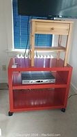 Red two-level rolling table with a DVD player on the middle shelf, solid wood side table with drawer placed on top.