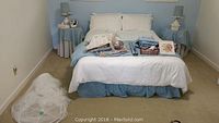 Full bed view showing white base bedding with blue bed skirt, two blue fabric-covered side tables with matching lamps, and bedding accessories on bed.