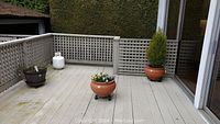 View of four planter pots on outdoor patio deck including a round clay pot with cedar, a square red clay pot on wheels, a wine barrel style resin planter, and another clay pot.