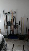 Wide view of all garden tools lined up against a wall showing long handles and variety of heads including shovels, rakes and brooms.